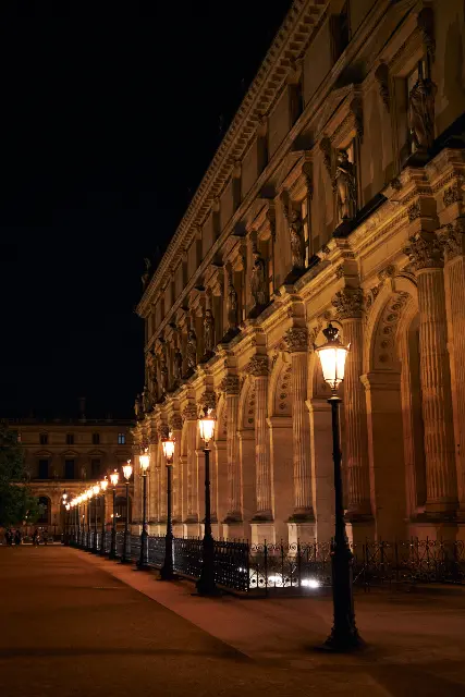 Ornate classical architecture lines a dark walkway under the warm glow of vintage street lamps.