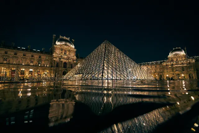 The illuminated Louvre Pyramid stands in the courtyard of the historic palace under a dark night sky.