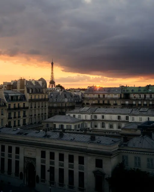 Eiffel Tower rises above Parisian rooftops under a dramatic sunset sky.