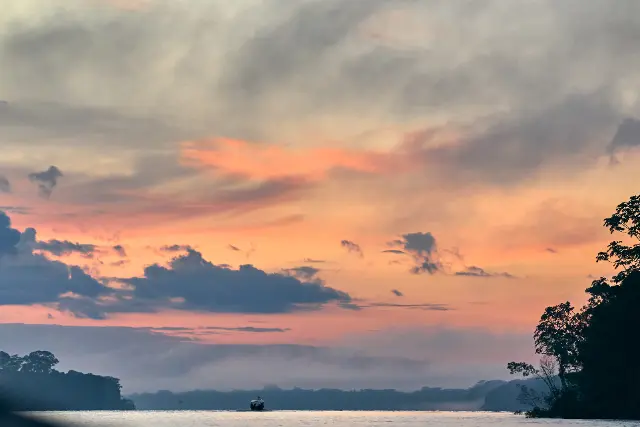 A small boat floats on a calm lake under a vast, cloudy sky during a soft sunset.