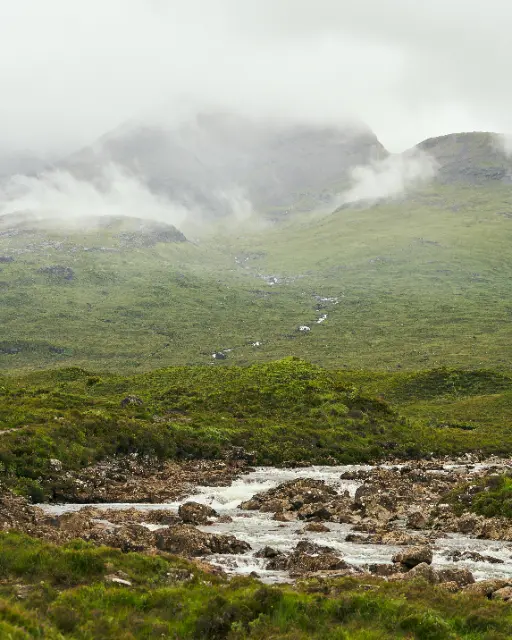 Mist-covered mountains rise above a lush green valley where a small stream flows through rocky terrain.