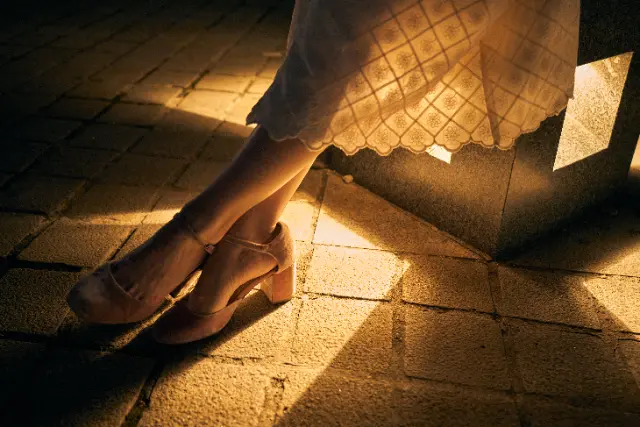 A person's legs and lace-hemmed skirt rest on a stone bench under warm, low-angled sunlight.