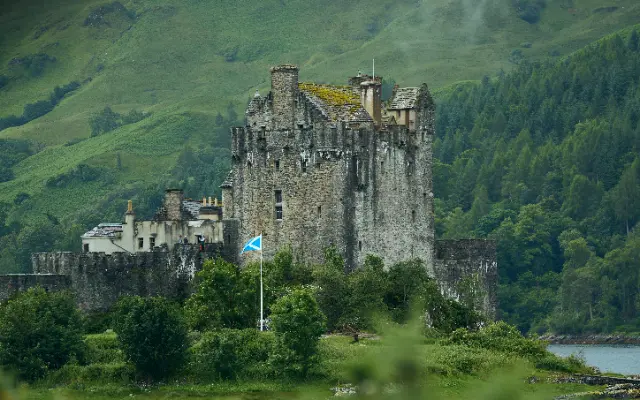 An ancient stone castle stands prominently against a backdrop of lush green mountains under soft, diffused daylight.