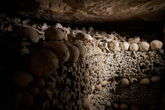 Stacked human skulls and bones fill a dark, subterranean ossuary illuminated by a single, dramatic light source.