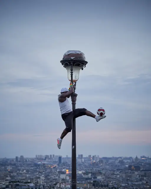 A man performs a trick on a tall street lamp overlooking a sprawling cityscape under a dim twilight sky.