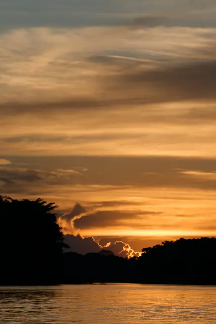Silhouetted trees line a calm river under a vast, cloud-filled sky during a golden sunset.
