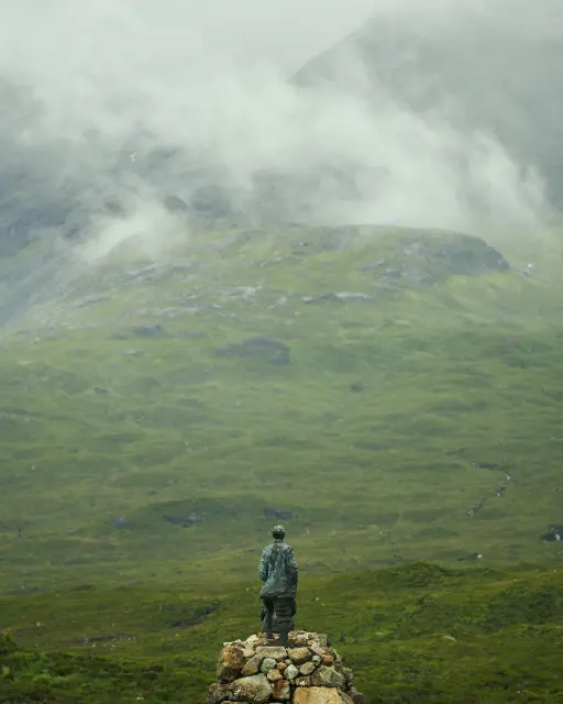 A lone figure stands atop a stone cairn overlooking a vast, misty mountain valley under soft, diffused light.