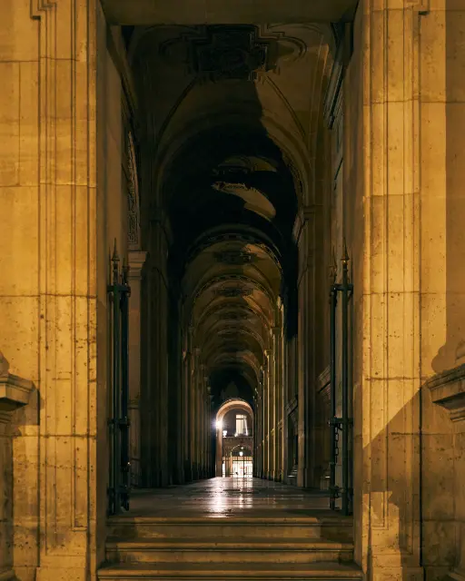 Tall stone pillars frame a long, vaulted arcade illuminated by dim, warm artificial light.